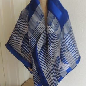 May include: A square silk scarf with a geometric pattern in shades of blue and gray. The scarf is displayed on a mannequin head, showcasing its design and drape. The scarf's edges are a vibrant blue.