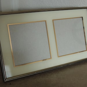 May include: A rectangular picture frame with a metallic finish and a textured outer edge. The frame features a cream-colored mat board with two square openings, each bordered by a thin gold-toned trim, designed to hold two separate photographs.