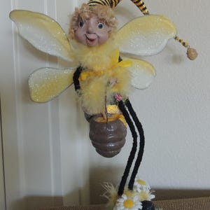 May include: A bee-themed figurine featuring a character with curly blonde hair, wearing a yellow and black striped hat and fuzzy yellow body. It has glittery white wings, long black pipe-cleaner legs, and holds a brown honey pot over white daisies.