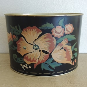 May include: Black metal planter with a floral design featuring orange, yellow, and green flowers. The planter has a gold rim and a black and white striped band around the bottom.