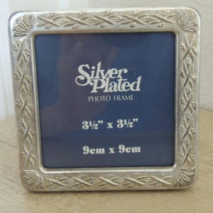 May include: A square silver-plated photo frame with an embossed floral border. The dark blue insert features the white text Silver Plated PHOTO FRAME, 3 1/2" x 3 1/2", and 9cm x 9cm. The frame has rounded corners and a metallic finish.
