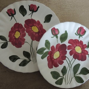 May include: Two white ceramic plates with a floral design. The plates feature red and green flowers with yellow centers. The plates are slightly scalloped on the edges.