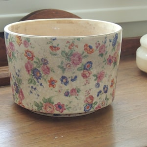 May include: A white ceramic planter with a floral pattern in shades of pink, blue, and orange. The planter has a round shape and a smooth, glossy finish.