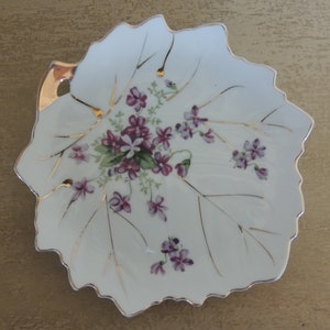 May include: A white porcelain leaf-shaped dish with a gold rim and a floral design of purple and green flowers. The dish has a small gold handle on the side.