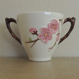 May include: A white ceramic creamer with brown handles and a pink floral design. The creamer features a raised design of a branch with pink flowers and green leaves.