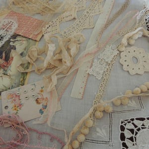 May include: A collection of cream and pink lace trims, ribbons, and fabric scraps arranged with a vintage postcard featuring two children, a small card with four white buttons and a cherub illustration, and a string of small cream pom-poms.