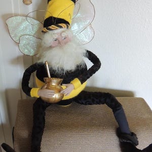 May include: A decorative doll dressed in a yellow and black striped bee costume with a matching pointed hat, white beard, and iridescent wings. The figure holds a small gold-colored vessel with a thin straw and wears black patterned pants.