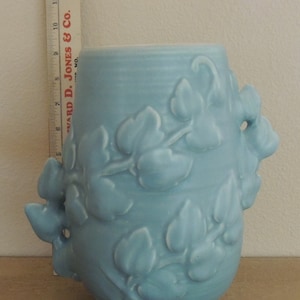 May include: A light blue ceramic vase with a floral design. The vase has two handles that are shaped like leaves. The vase is about 6 inches tall.