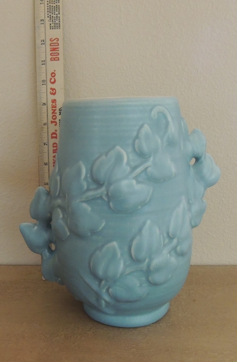 May include: A light blue ceramic vase with a floral design. The vase has two handles that are shaped like leaves. The vase is about 6 inches tall.