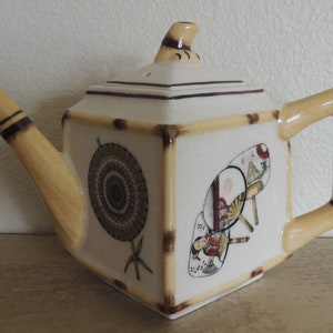 May include: A white ceramic teapot with a bamboo-style handle and lid. The teapot is decorated with a Japanese-inspired design featuring fans and a woman in traditional clothing.