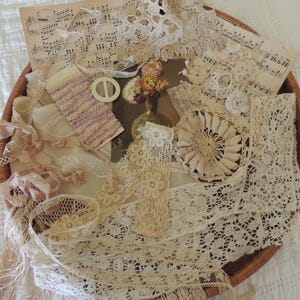 May include: A collection of cream and off-white lace trims, ribbons, and fabric scraps arranged on a round woven tray. The assortment includes floral lace patterns, a small white circular buckle, a piece of vintage sheet music, and a floral print.