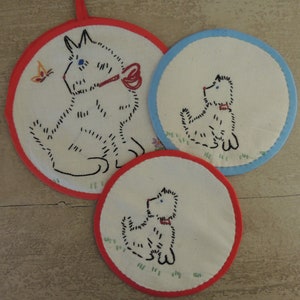 May include: Three embroidered white cotton pot holders with red and blue trim. Each pot holder features a black embroidered image of a dog.