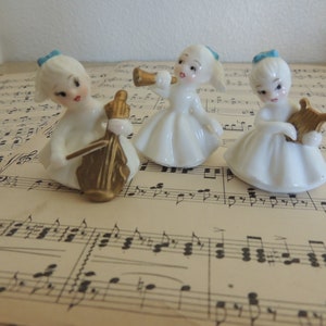 May include: Three white porcelain figurines of angels playing musical instruments. The angel on the left is playing a cello, the angel in the middle is playing a trumpet, and the angel on the right is playing a harp. All three angels have blue bows in their hair.