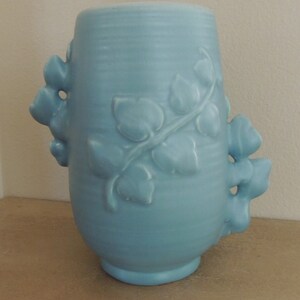 May include: A light blue ceramic vase with a vine and leaf design. The vase has two handles that are shaped like leaves.