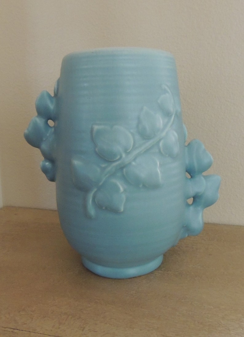 May include: A light blue ceramic vase with a vine and leaf design. The vase has two handles that are shaped like leaves.