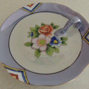 May include: A vintage ceramic serving dish with a handle. The dish features a light purple rim with geometric designs in yellow, blue, and red. The center has a floral arrangement of pink, orange, and blue flowers with green leaves.