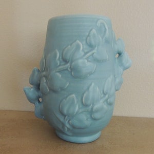 May include: A light blue ceramic vase with a raised ivy design. The vase has a rounded body and a wide mouth.