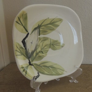 May include: A square, off-white ceramic plate with rounded corners, featuring a hand-painted floral design. The design includes green leaves and a white flower bud on a black stem. The plate is displayed on a clear plastic stand.