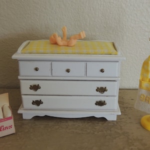 May include: A white dresser with four drawers and a yellow and white checkered changing pad on top. A small plastic doll is lying on the changing pad. A yellow and white checkered baby bag hangs on a white stand with a yellow clown doll on top.