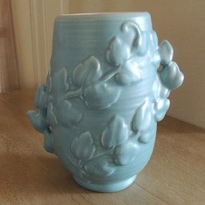 May include: A light blue ceramic vase with a floral design. The vase has a rounded body and a wide mouth. The floral design is embossed and features leaves and flowers.