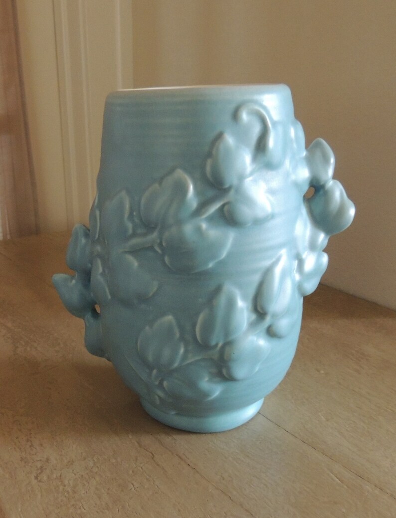 May include: A light blue ceramic vase with a floral design. The vase has a rounded body and a wide mouth. The floral design is embossed and features leaves and flowers.