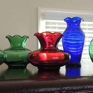 May include: Five glass vases in different colors and shapes. Two green vases with ruffled tops, one red vase with a ruffled top, one blue vase with a ruffled top, and one round green vase.