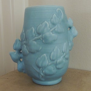 May include: A light blue ceramic vase with a floral design. The vase has a rounded body and a wide mouth. The floral design is embossed and features leaves and flowers.