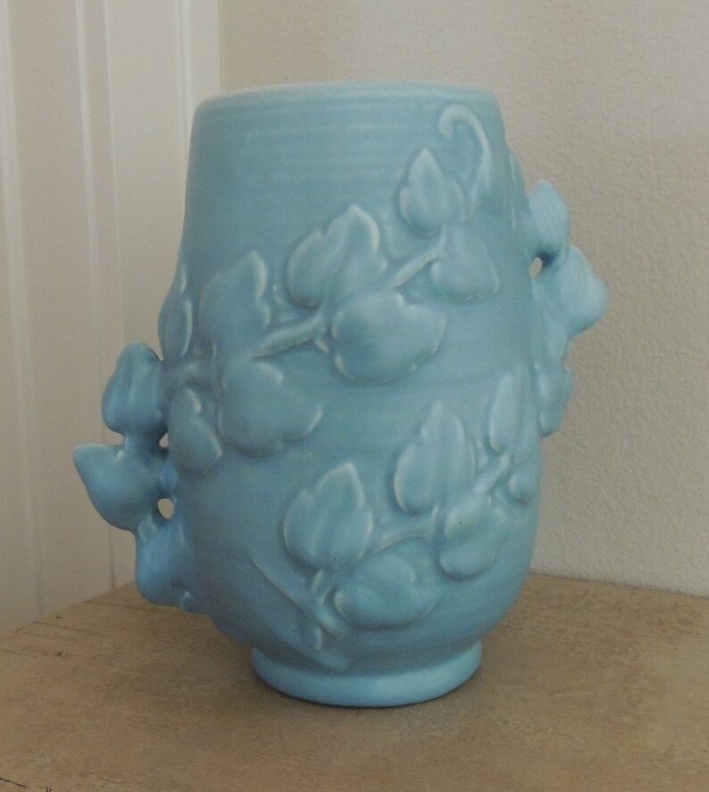 May include: A light blue ceramic vase with a floral design. The vase has a rounded body and a wide mouth. The floral design is embossed and features leaves and flowers.