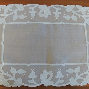 May include: A rectangular, white, embroidered doily with a floral design. The edges are scalloped, and the embroidery features roses and leaves. The doily is made of a sheer fabric and is suitable for decorative purposes.