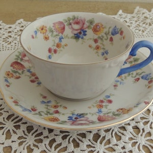 May include: A white teacup and saucer with a floral pattern in shades of pink, yellow, blue, and green. The cup has a blue handle and a gold rim. The saucer also has a gold rim.