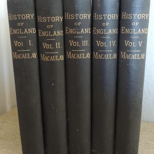 May include: A collection of five antique books titled "History of England" by Macaulay. The spines are dark gray with gold lettering, displaying the volume number and author's name. The books are arranged side by side.
