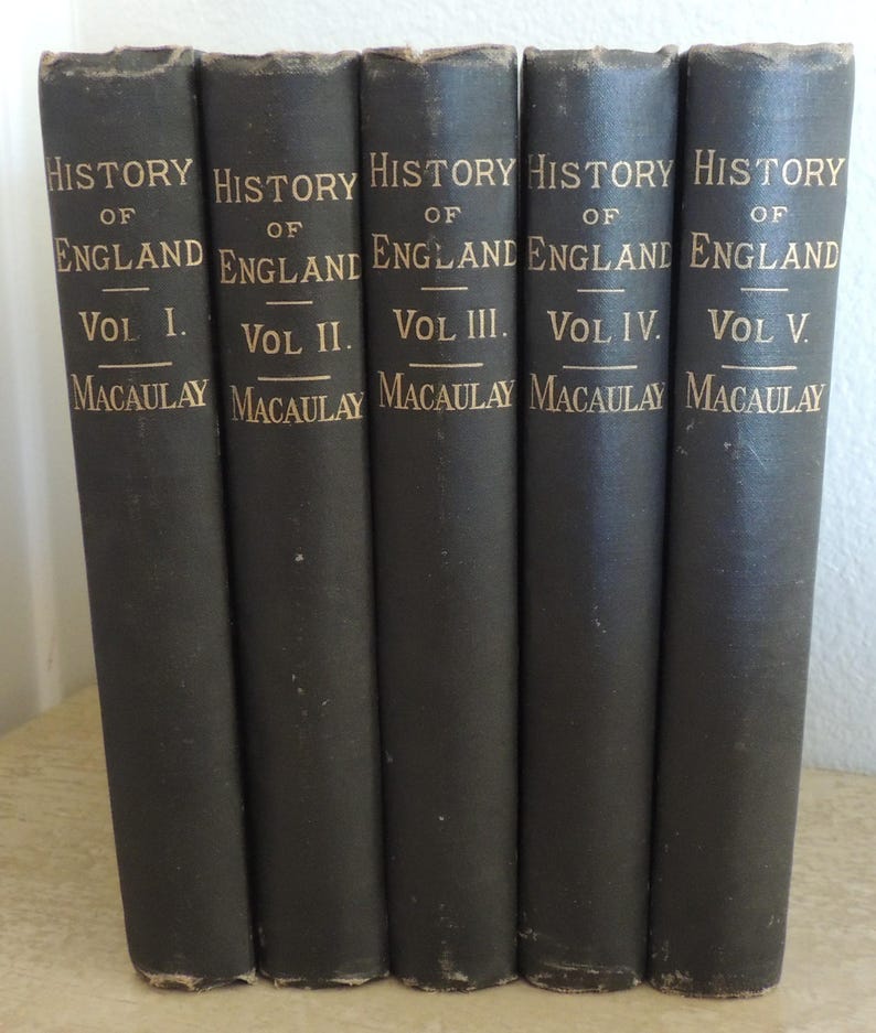 May include: A collection of five antique books titled "History of England" by Macaulay. The spines are dark gray with gold lettering, displaying the volume number and author's name. The books are arranged side by side.