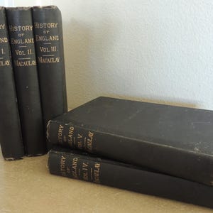May include: A collection of antique books with black covers and gold lettering. The spines read "History of England" and "Macaulay," with volume numbers I, II, and III visible. Two additional volumes are stacked in front, with volumes IV and V visible.