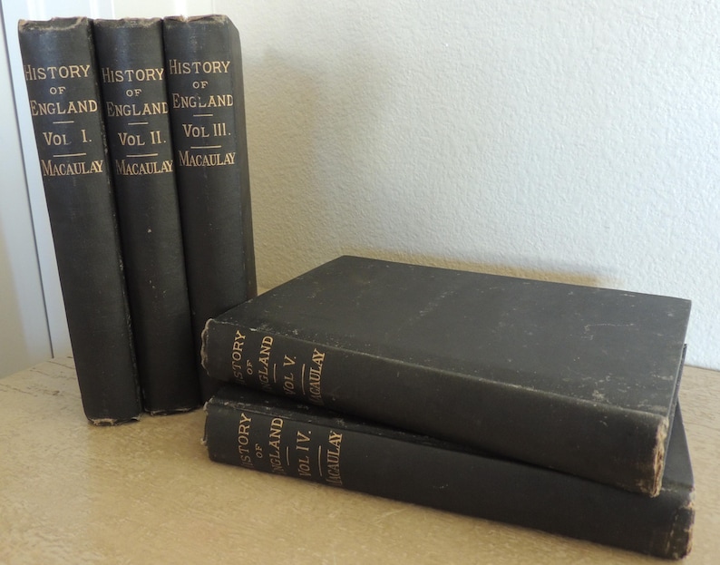 May include: A collection of antique books with black covers and gold lettering. The spines read "History of England" and "Macaulay," with volume numbers I, II, and III visible. Two additional volumes are stacked in front, with volumes IV and V visible.
