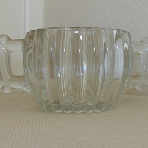 May include: A clear glass sugar bowl with two handles. The bowl has vertical ridges and a rounded bottom.