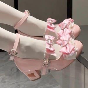May include: Pink patent leather Mary Jane shoes with ankle straps and heart-shaped cutouts on the heels. The shoes feature satin bows, rhinestone buckles, and lace trim, creating a whimsical and feminine design.