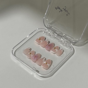 May include: A set of press-on nails in a clear plastic case. The nails have a light pink base with white tips, gold accents, and a dotted design. Some nails feature pink floral details.