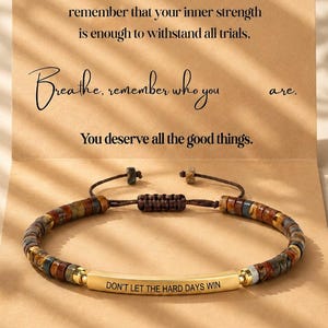 Può includere: Un braccialetto di perline con una barra di metallo dorato incisa con il testo "DON'T LET THE HARD DAYS WIN" presentato su un cartoncino color sabbia con il messaggio stampato: "Life may be harsh at times, but always remember that your inner strength is enough to withstand all trials. Breathe, remember who you are. You deserve all the good things."