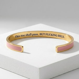 May include: A gold-tone metal cuff bracelet featuring a pink enamel inlay along the outer band. The interior surface is engraved with the black text, "This too shall pass, BUT FUCKING HELL". The bracelet has an open-ended design for adjustment.