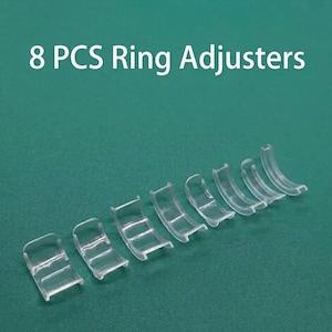Puede incluir: Ocho ajustadores de anillos de plástico transparente se muestran sobre una superficie verde. El texto "8 PCS Ring Adjusters" se muestra en blanco en la parte superior. Los ajustadores son curvos y están diseñados para reducir el tamaño de un anillo.