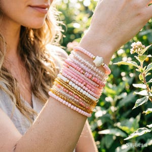 Op de afbeelding: Een verzameling kralenarmbanden in roze, goud en heldere tinten, gedragen om een pols. De armbanden bevatten verschillende kralenmaten en materialen, waaronder een armband met een schelpenbedel. De armbanden zijn een mix van elastiek en draad.