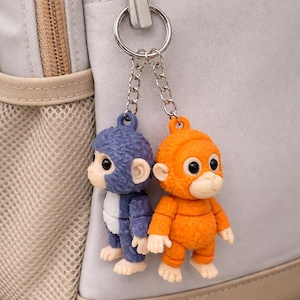 Può includere: Un portachiavi con due piccole statuette a forma di scimmia. Una è blu e bianca, l'altra arancione. Sono attaccate a una catena e un anello argentati, appesi a uno zaino grigio chiaro.