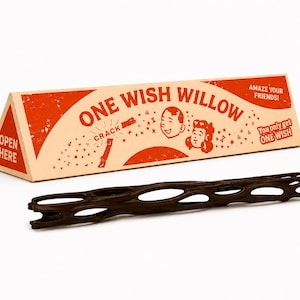 Könnte beinhalten: Eine dreieckige Pappschachtel mit orangefarbenen und cremefarbenen Grafiken, beschriftet mit ONE WISH WILLOW, AMAZE YOUR FRIENDS und YOU ONLY GET ONE WISH, sowie ein langes, dunkelbraunes, zylindrisches Holzobjekt mit poröser Gitterstruktur.