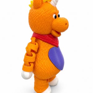 Peut inclure: Une figurine de licorne orange texturée avec une crinière jaune, une corne blanche et un bandana rouge autour du cou. La figurine présente un écusson ovale violet sur la poitrine, des pattes blanches et un bras articulé avec des pièces en plastique orange.