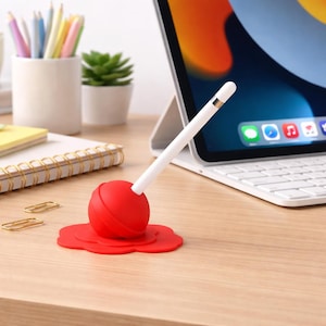 Puede incluir: Un soporte de lápiz de silicona roja con forma de piruleta, que sostiene un lápiz blanco. El soporte está sobre un escritorio de madera junto a una tableta y otros útiles de oficina. El soporte tiene unos 5 cm de diámetro.