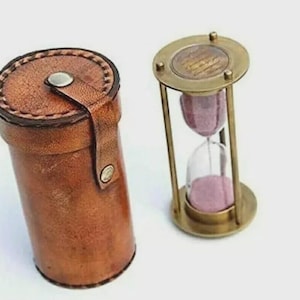 May include: A vintage-style hourglass with a brass frame and pink sand, next to a brown leather cylindrical case. The hourglass is a classic timekeeping device, and the case has a snap closure.