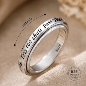 Puede incluir: Un anillo de plata con la inscripción "This too shall Pass" y una banda exterior giratoria. El anillo está marcado "S925" y "925 Sterling Silver". El anillo tiene un diseño simple y elegante.