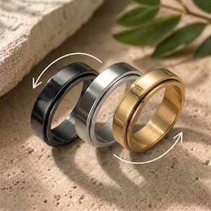 Könnte beinhalten: Drei Metall-Spinner-Ringe in Schwarz, Silber und Gold. Die Ringe haben ein schlichtes, modernes Design mit einem rotierenden Außenband. Der goldene Ring hat ein rotbraunes Innenband. Die Ringe werden auf einer strukturierten Oberfläche mit einem grünen Blatt im Hintergrund präsentiert.