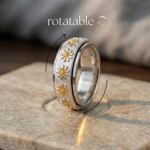 Könnte beinhalten: Ein silberfarbener Ring mit matter Oberfläche und einem wiederkehrenden Muster aus kleinen, goldfarbenen Gänseblümchen rund um das Band. Über dem Ring steht das Wort rotatable, ergänzt durch zwei gebogene Pfeile, die eine Drehbewegung anzeigen.
