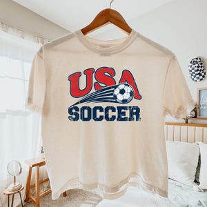 May include: A cream-colored short-sleeve t-shirt featuring a graphic print with the red text USA above a blue soccer ball in motion, followed by the blue text SOCCER. The design has a distressed, weathered texture throughout the lettering.